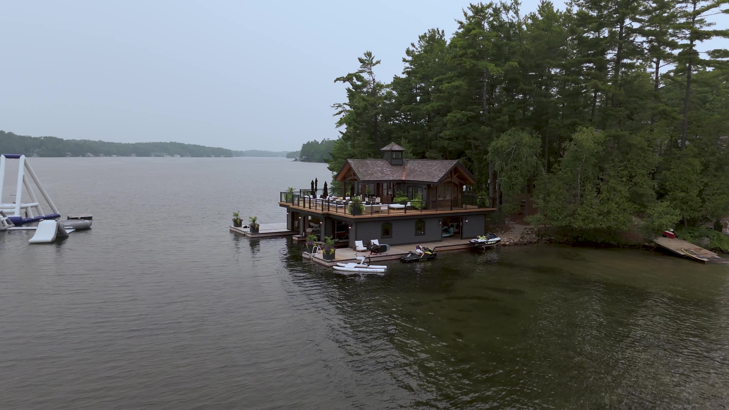Robert Herjavec and Kym Johnson-Herjavec’s two-storey Muskoka boathouse with an upper deck, docks and watercraft along a forested Ontario shoreline.